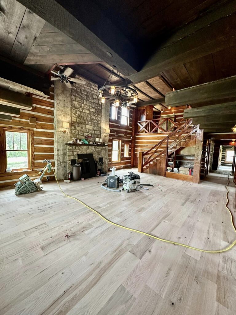 A rustic cabin interior featuring newly installed light hardwood floors, a large stone fireplace, exposed wooden beams, and a wooden staircase leading to a loft area. The room is partially set up with construction equipment, indicating ongoing work.